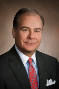 A professional man in a dark suit with a white shirt and red tie is posing against a neutral brown background. He has short hair and a light complexion, and he appears to be smiling slightly.