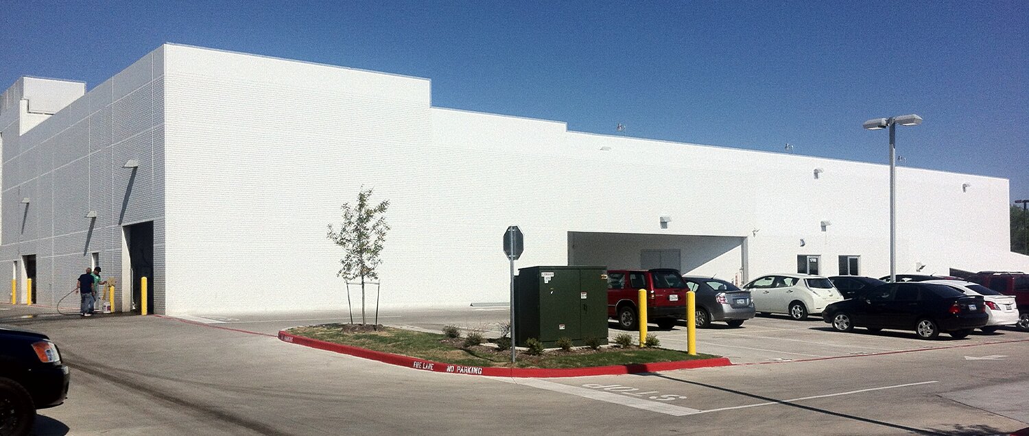 Nissan1 A large, white industrial building with a flat roof under a clear blue sky. Several cars are parked in the adjacent lot. A few small trees and a traffic sign are visible near the entrance.
