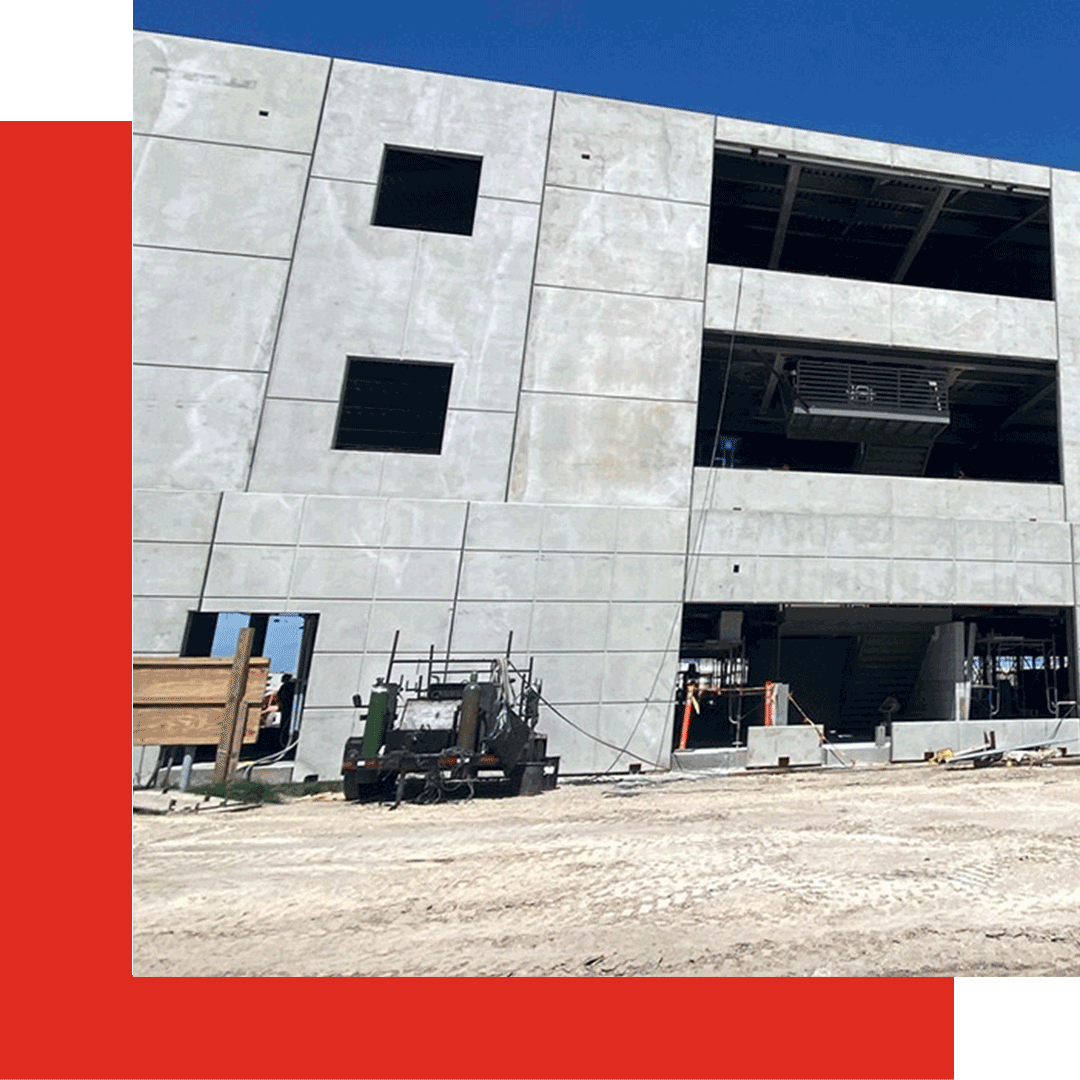 A partially constructed building with exposed concrete walls and large window openings. Two construction vehicles are parked in front on a dirt surface. The sky is clear and blue.