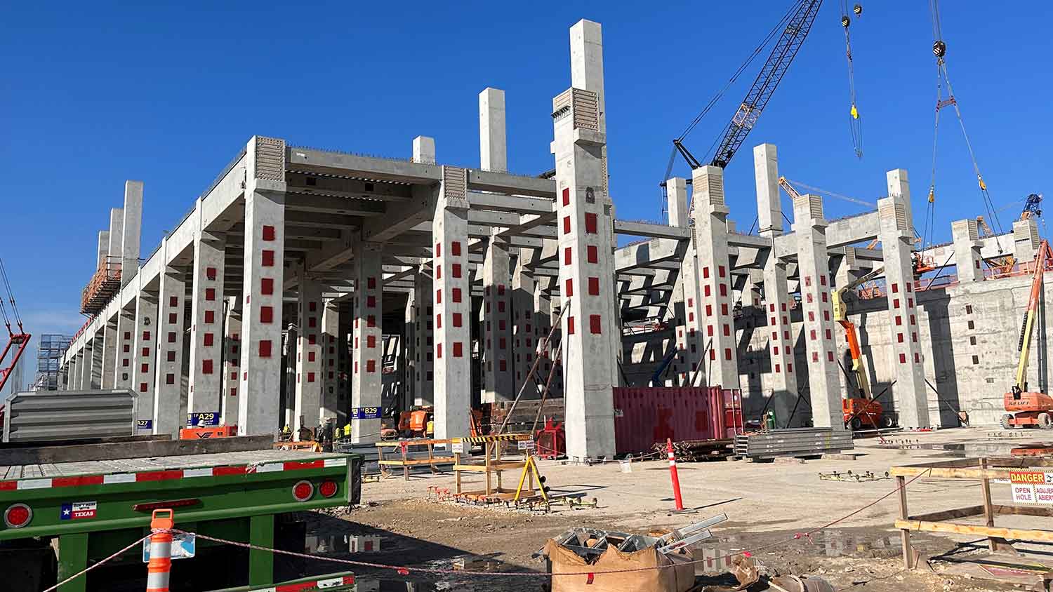 A large concrete structure under construction, with columns and beams visible. Construction equipment, materials, and cranes surround the building site on a clear, sunny day.