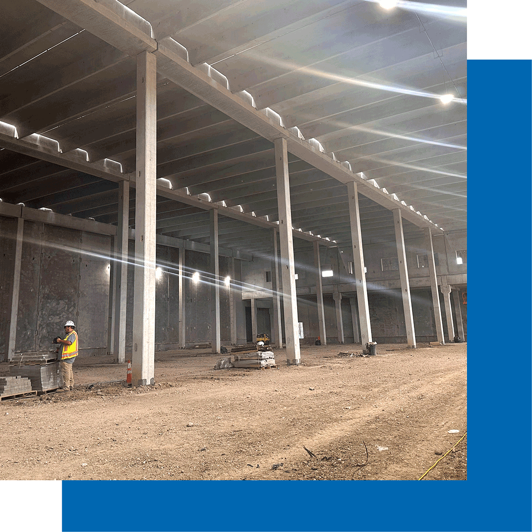 A large, unfinished warehouse with tall concrete columns and beams. Two construction workers in safety vests and helmets stand on the dirt floor surrounded by building materials. Sunlight streams through the ceiling.