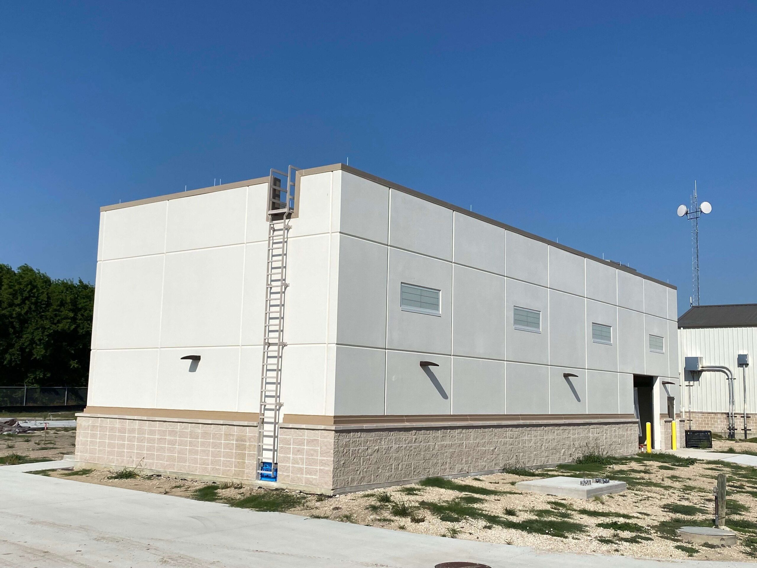 A rectangular, windowless, beige utility building with a ladder attached to one side is shown under a clear blue sky. The building sits on a patch of sparse grass and dirt next to a paved area.