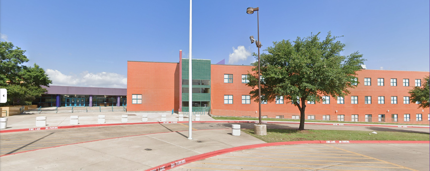 A large, modern brick school building with blue doors, green glass accents, and multiple windows, set behind a flagpole, parking lot, and trees under a clear blue sky.