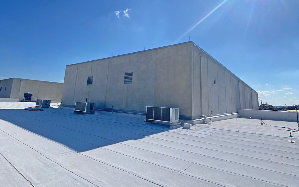 A large, flat commercial building roof with HVAC units and vents, covered in bright white roofing material under a clear blue sky with sunlight casting sharp shadows.