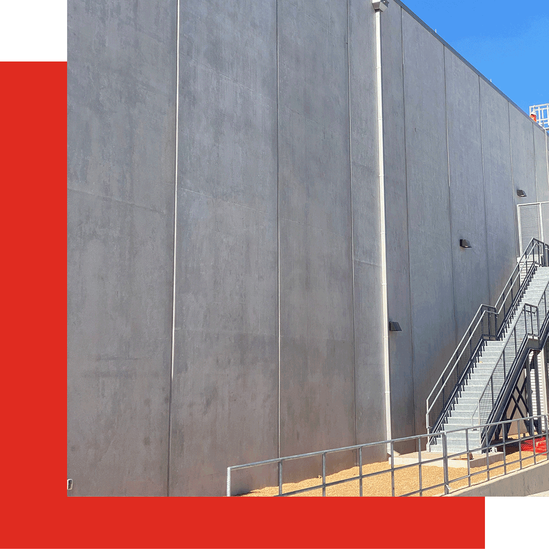 A large gray concrete building wall with a black metal staircase and railing on the right side, set against a bright blue sky. Bold red and white geometric shapes frame the left and bottom edges.
