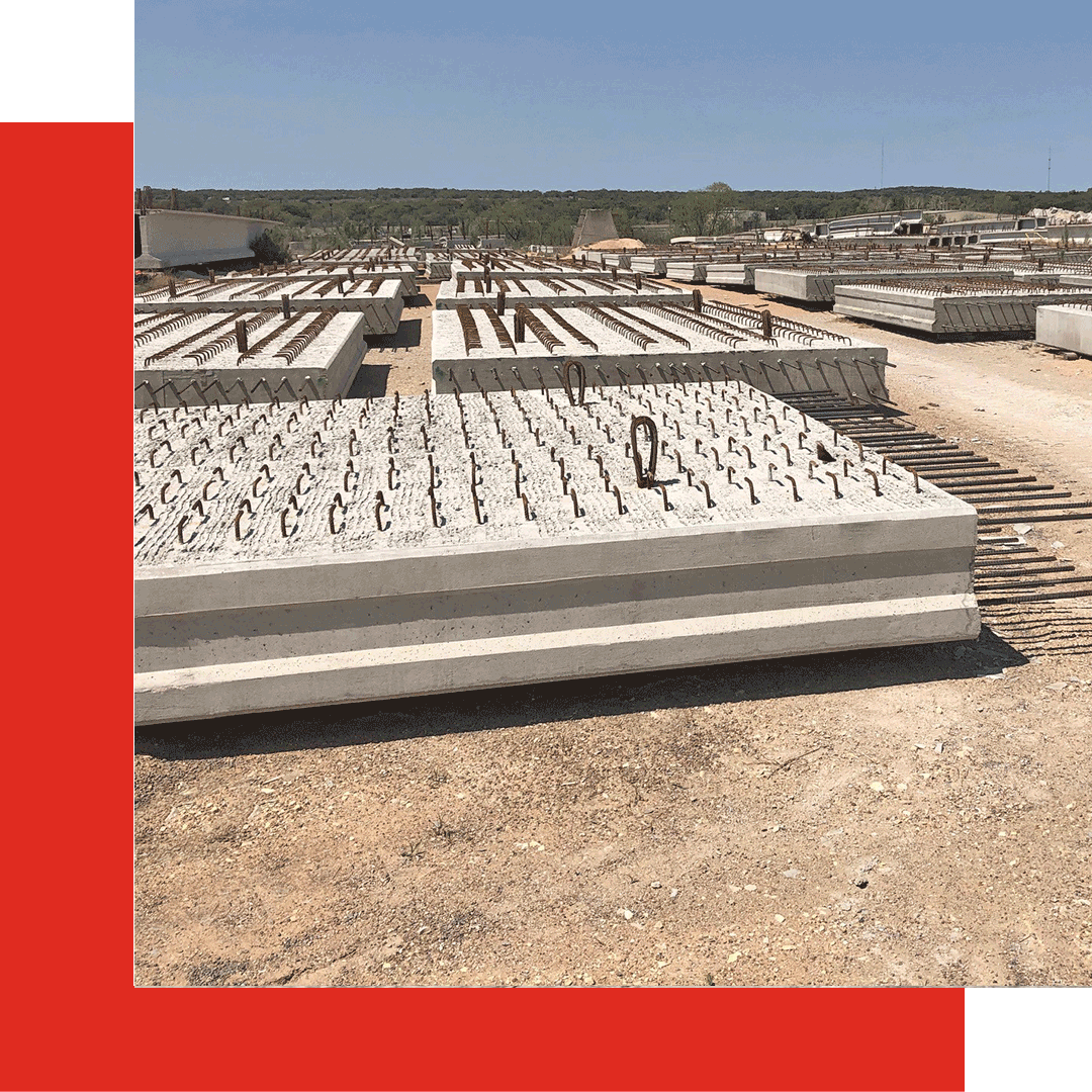 Large concrete slabs with metal rebar protruding are stacked outdoors on a sandy lot under a clear blue sky. Rows of similar slabs extend into the distance. Red geometric shapes frame the image's left and bottom edges.