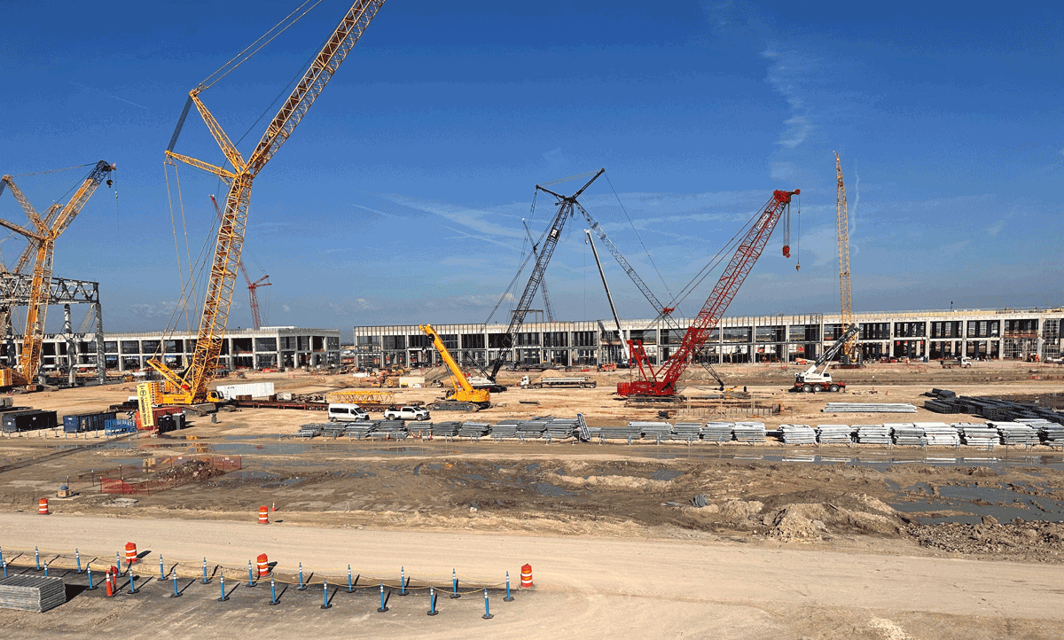 Large construction site with several tall cranes, construction vehicles, and building materials scattered around. A partially built structure is visible in the background under a clear blue sky.