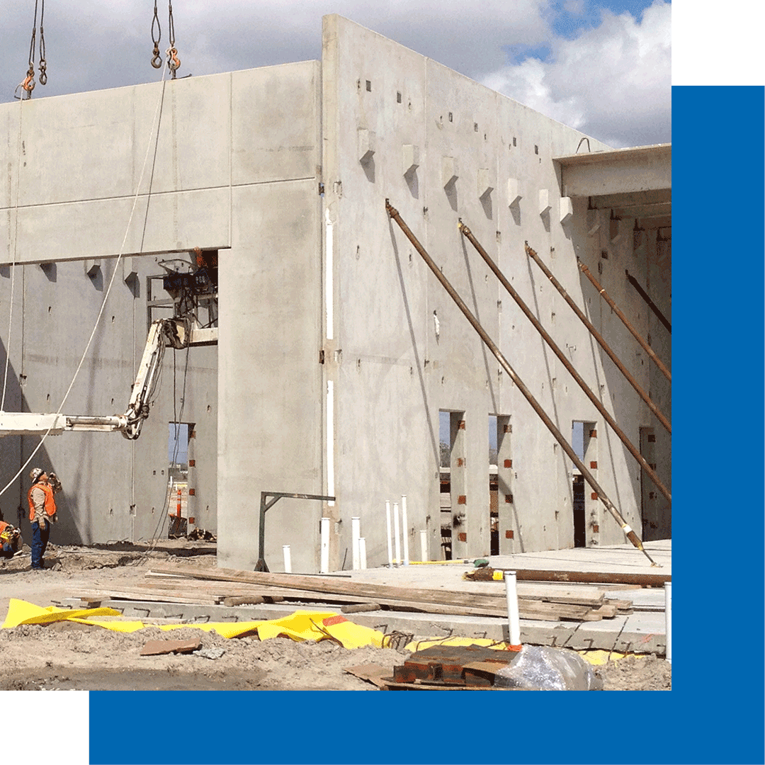 Construction workers operate a boom lift near a large concrete building under construction, with support braces holding up tall concrete walls and construction materials scattered on the ground.