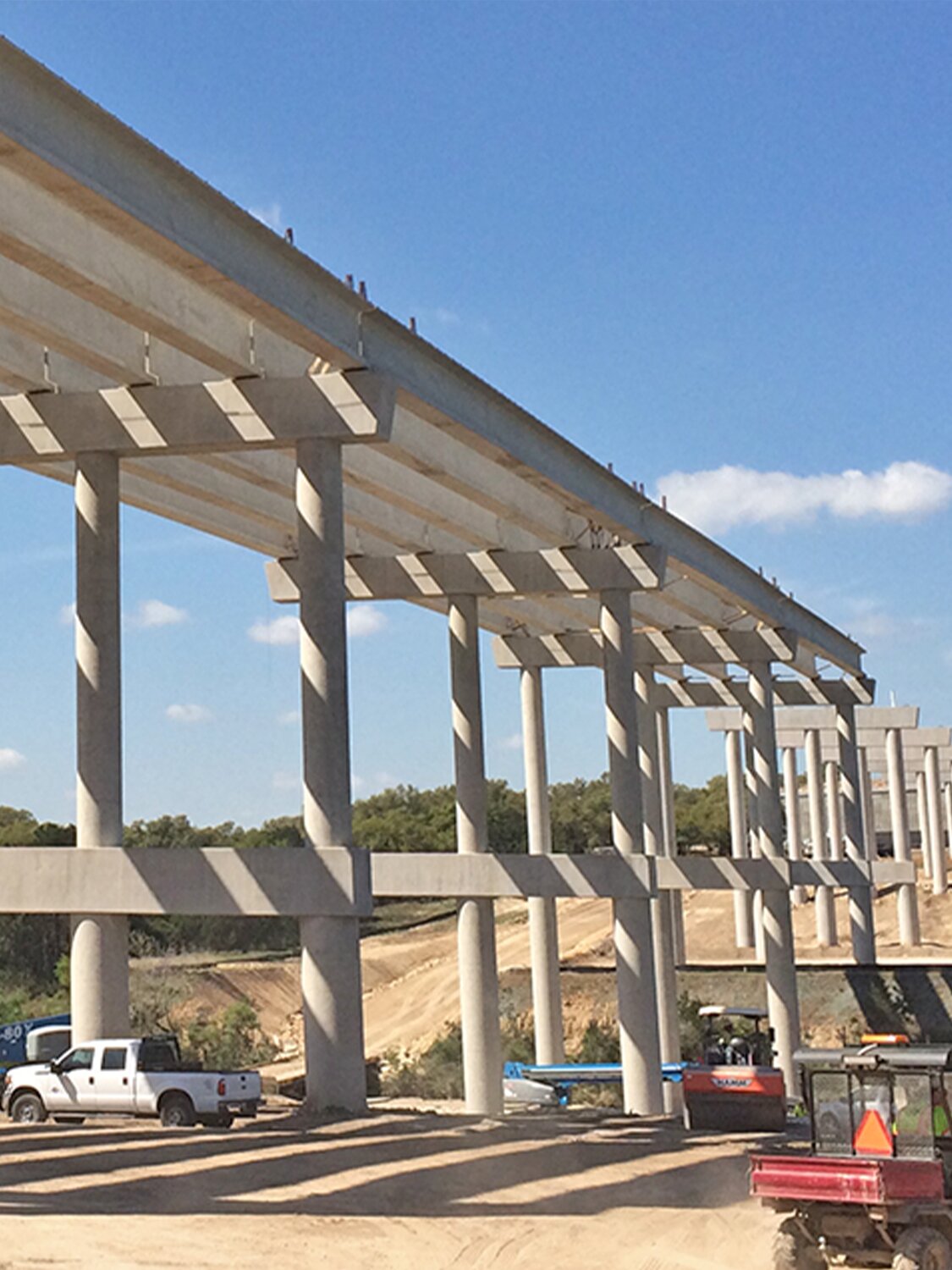 Concrete bridge under construction with multiple support columns, vehicles, and construction equipment visible below, set against a blue sky with scattered clouds.