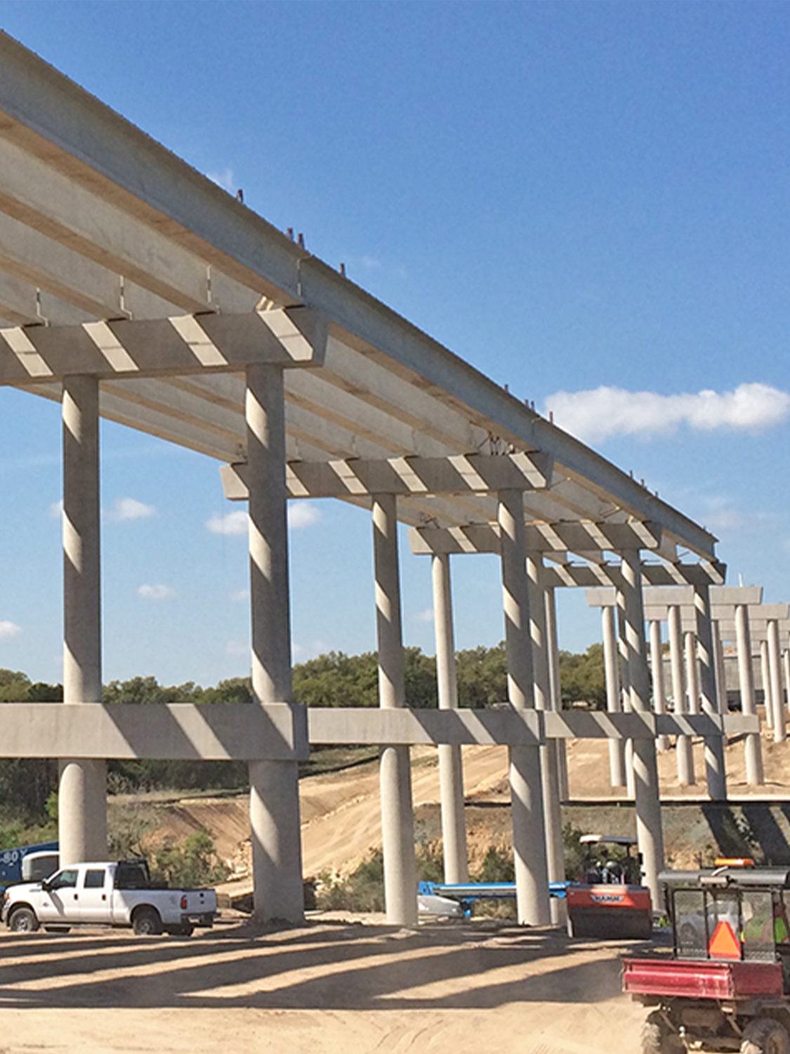 Concrete bridge under construction with multiple support columns, vehicles, and construction equipment visible below, set against a blue sky with scattered clouds.