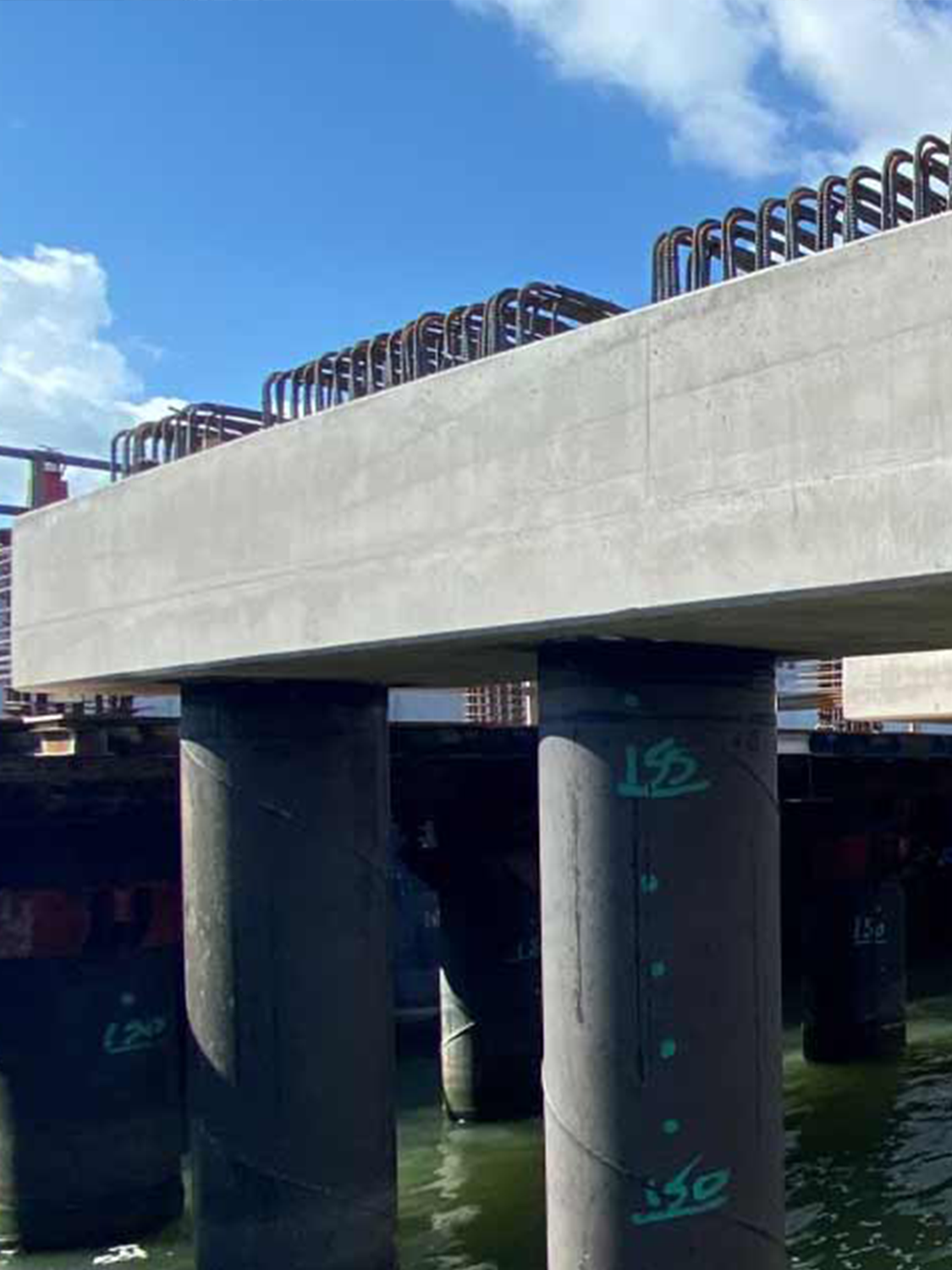 A concrete bridge under construction with exposed rebar on top, supported by large cylindrical pillars standing in water, under a partly cloudy blue sky.