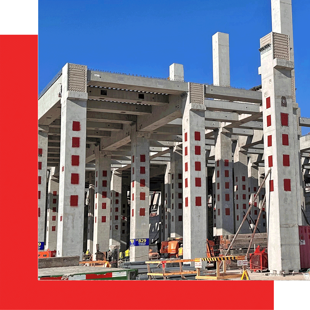 A concrete building under construction with tall columns marked by red squares; construction materials, barriers, and equipment are visible at the base under a clear blue sky.