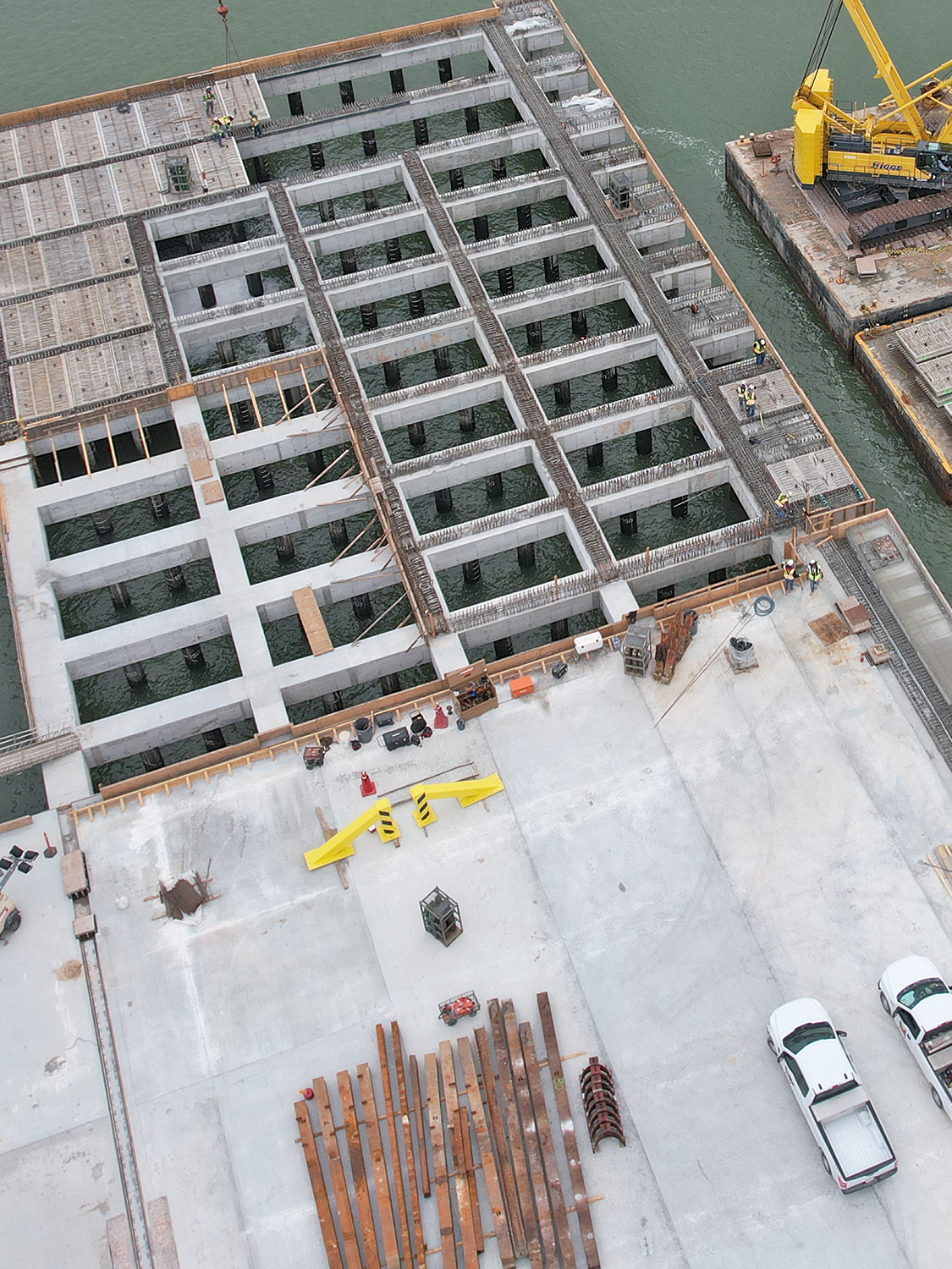DeckSlabs Aerial view of a large construction site by water, featuring a concrete foundation with a grid pattern, construction materials, two white vehicles, and a yellow crane on a barge.