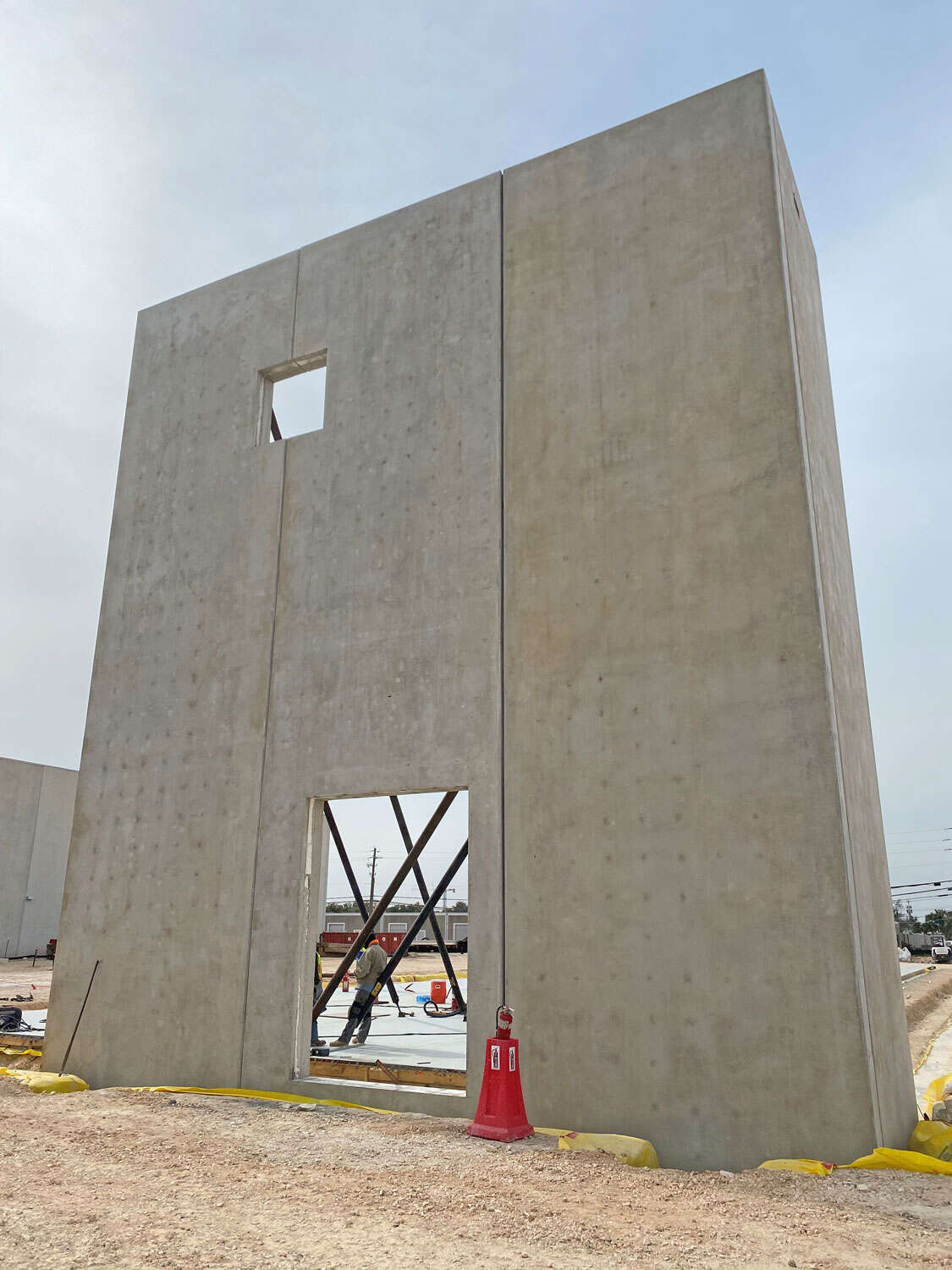 A tall concrete wall panel with a rectangular doorway and a small square window is supported by braces at a construction site. Workers and construction materials are visible behind the wall.