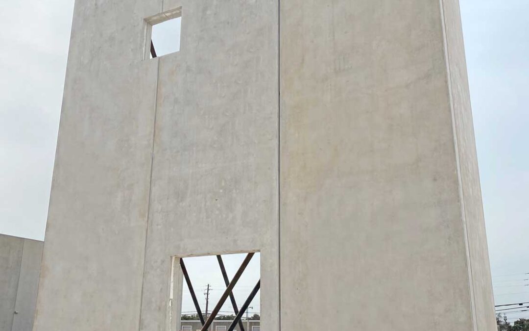 A tall, rectangular concrete wall with a door opening and a small window opening stands on a construction site. A worker and metal braces are visible through the door opening. The ground is bare dirt.