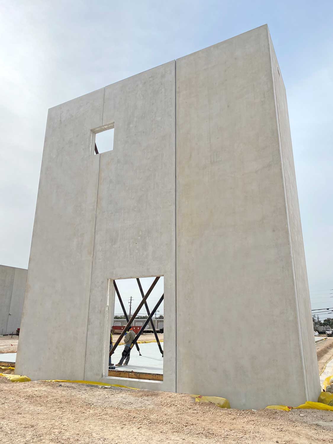 A tall, rectangular concrete wall with a door opening and a small window opening stands on a construction site. A worker and metal braces are visible through the door opening. The ground is bare dirt.