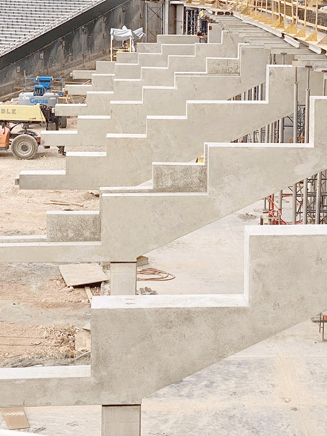 Rows of unfinished concrete stadium seating rise in stepped formation at a construction site, with scaffolding and equipment visible in the background.