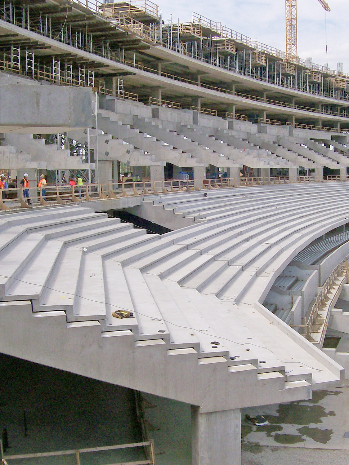 Rows of unfinished concrete stadium seating under construction, with workers in safety vests standing on the far side and scaffolding and cranes visible in the background.