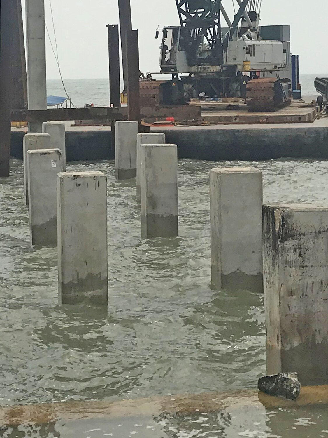 Concrete pilings are partially submerged in water at a construction site near the shore. In the background, a large crane sits on a barge, and the horizon is visible.