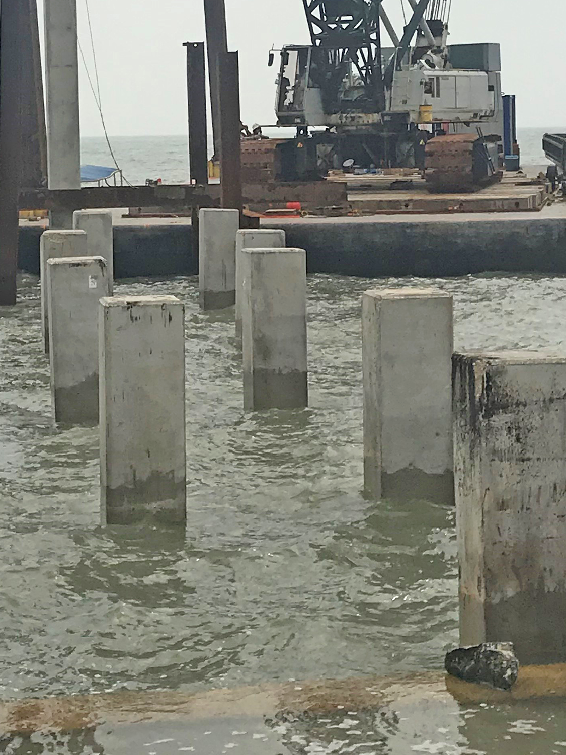 Concrete pilings are partially submerged in water at a construction site near the shore. In the background, a large crane sits on a barge, and the horizon is visible.