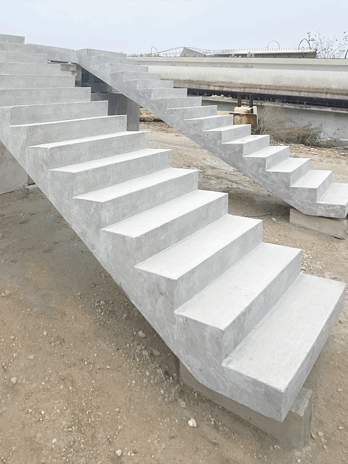 Two unfinished concrete staircases stand outdoors on a construction site with dirt ground; there are no railings, and construction materials and pipes are visible in the background.