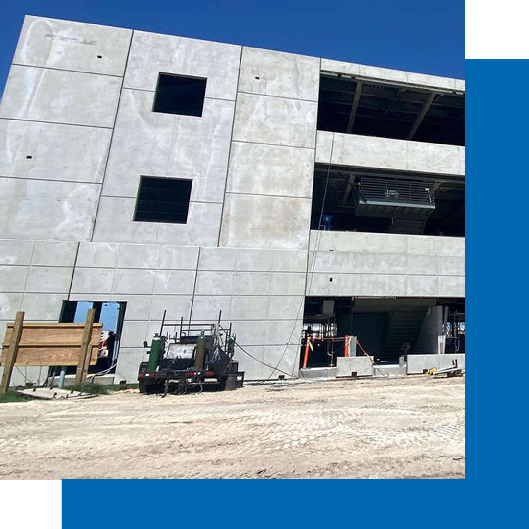 A large concrete building under construction, with unfinished windows and exposed floors, sits on a dirt lot under a clear blue sky. Construction equipment and materials are visible at ground level.