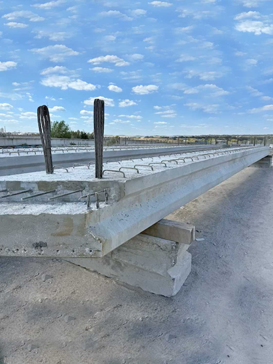 A concrete bridge beam with exposed steel reinforcement bars sits on a construction site under a bright blue sky with scattered clouds. The area is surrounded by dirt and distant trees.