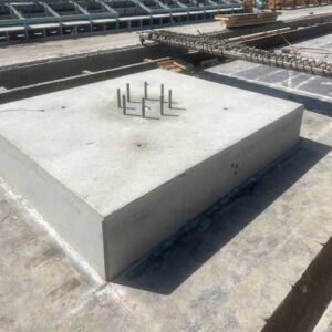 A large rectangular concrete foundation block with metal anchor bolts protruding from the top, situated on a construction site with building materials and scaffolding in the background.
