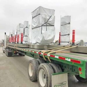 A flatbed truck carries several large objects covered in white protective wrapping and secured with straps. The objects are lined up and spaced apart, with red and white posts visible in between.