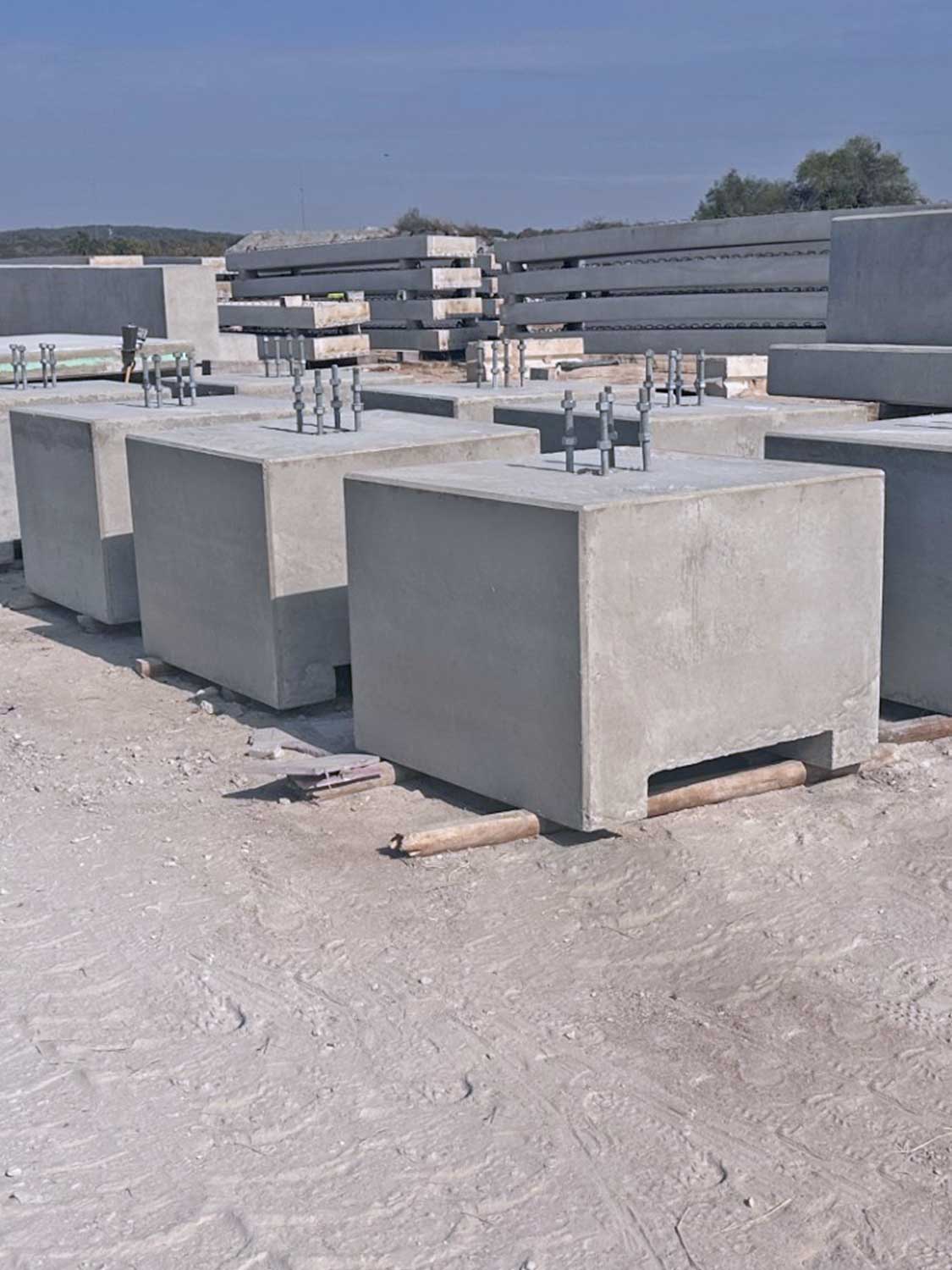 EquipmentPads Large concrete blocks with metal bolts on top are arranged on a dusty construction site. In the background, more concrete slabs and blocks are stacked. The area appears industrial and outdoors.