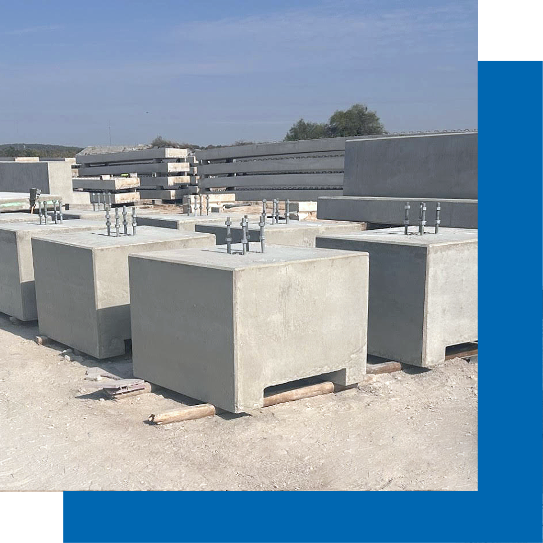 Large concrete blocks with metal bolts are stacked outdoors on a construction site, with more concrete slabs and beams visible in the background under a clear sky.