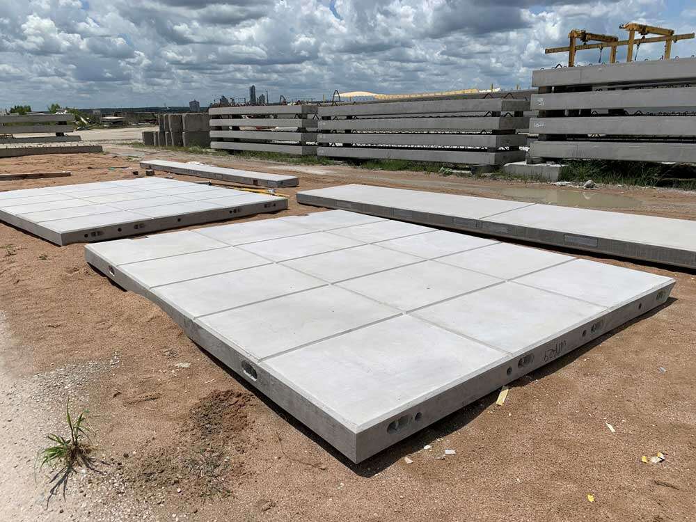 NAS1 Large precast concrete panels lie on sandy ground at an outdoor construction site, with stacks of additional concrete pieces and industrial equipment visible in the background under a partly cloudy sky.