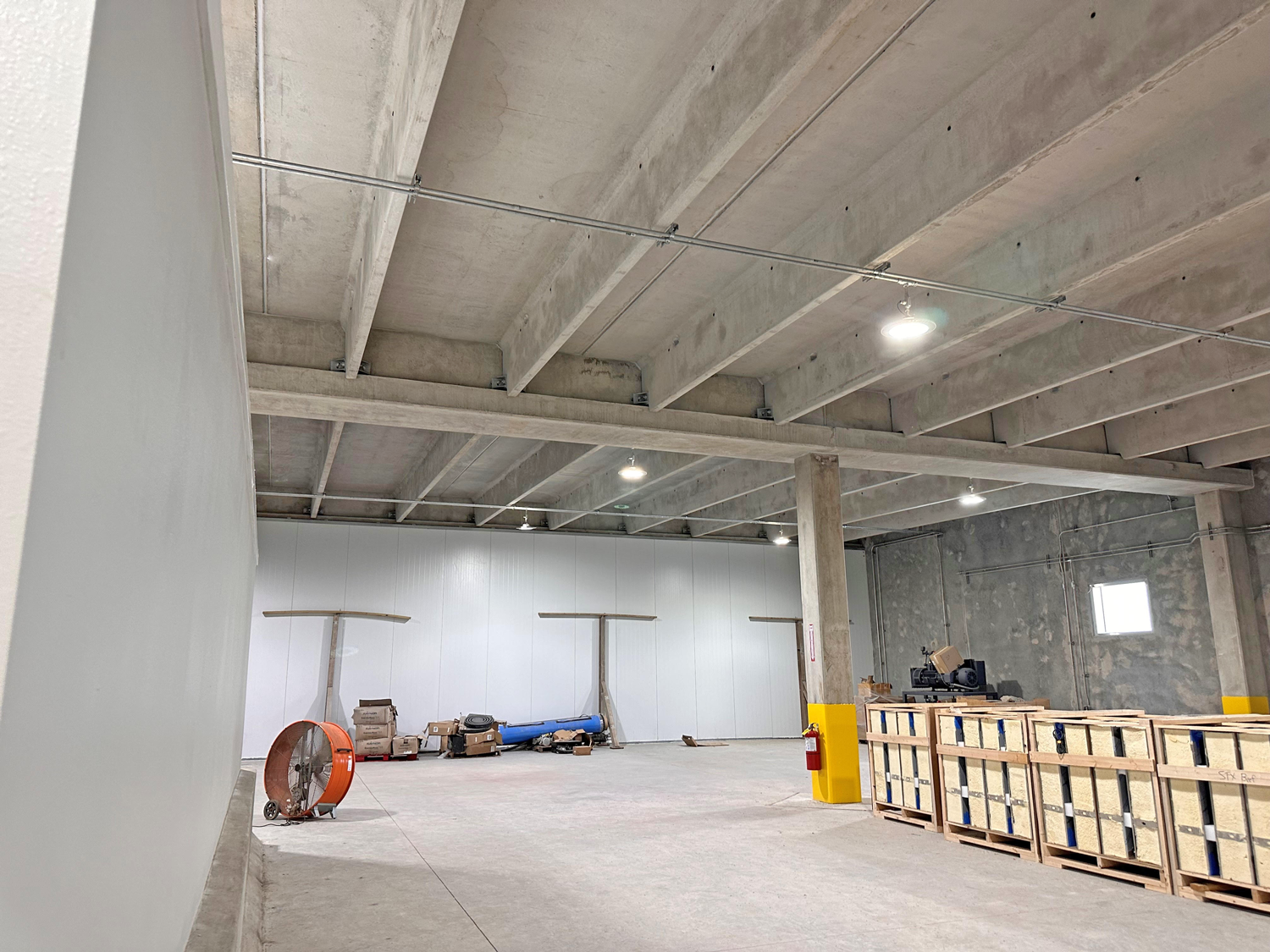 STX-2 A spacious industrial warehouse with concrete walls, exposed beams, and overhead lights. Crates, boxes, equipment, and a large orange fan are scattered around the unfinished, mostly empty interior.