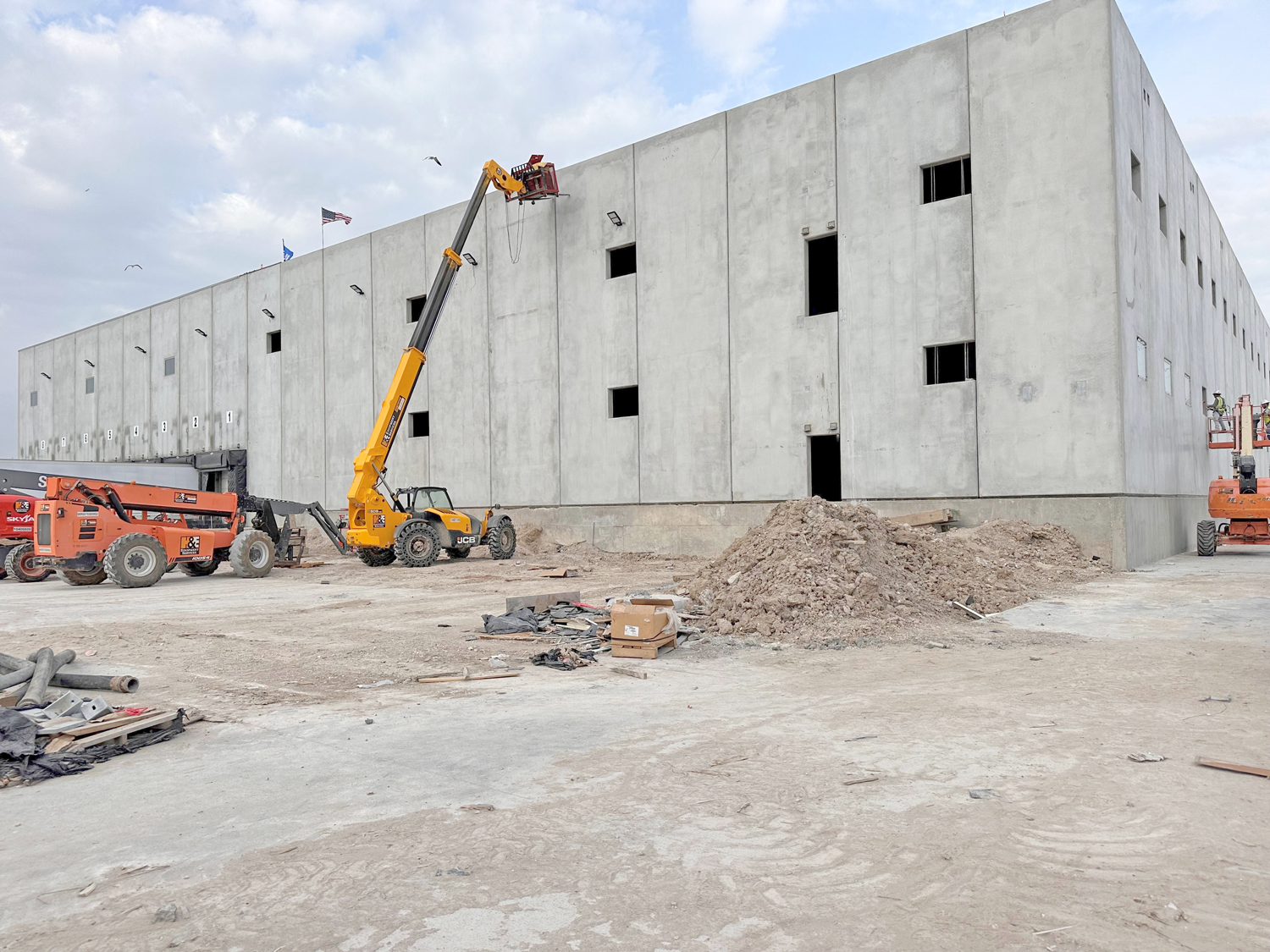 STX1 A construction site with a large, unfinished concrete building. A lift raises a worker near the top, while construction vehicles and piles of dirt are scattered across the ground. The sky is partly cloudy.