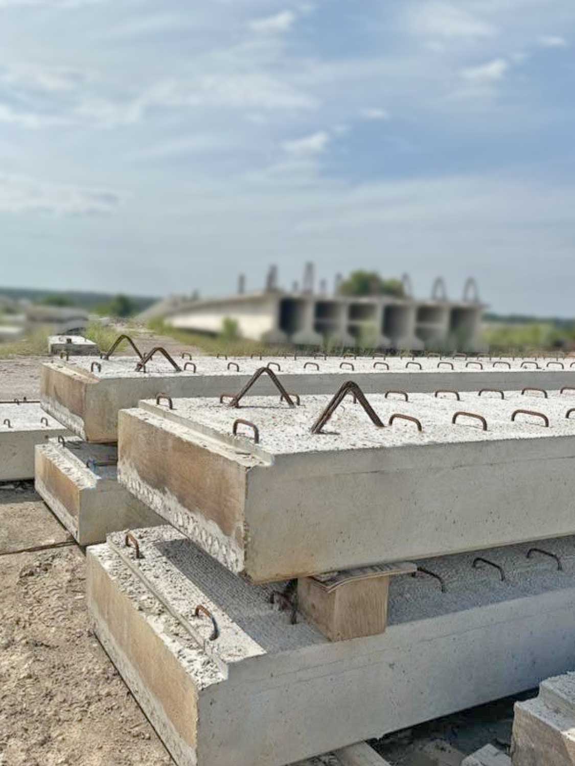 Stacks of large rectangular concrete slabs with metal loops sit on a construction site, with an unfinished building and blue sky in the background.