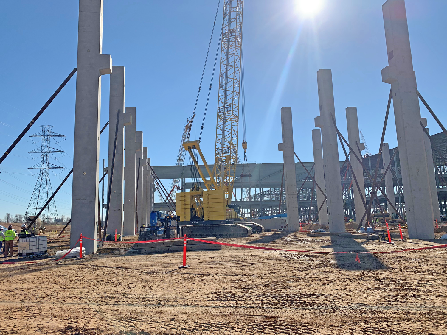 Tesla1 A construction site with tall concrete pillars, a yellow crane in the center, workers in safety vests, red caution tape, and a clear blue sky with the sun shining overhead.