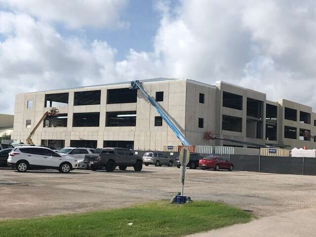 image001 (5) A large, unfinished concrete building under construction, with cranes and construction equipment on site. Several cars are parked in front of the building, and the sky is partly cloudy.