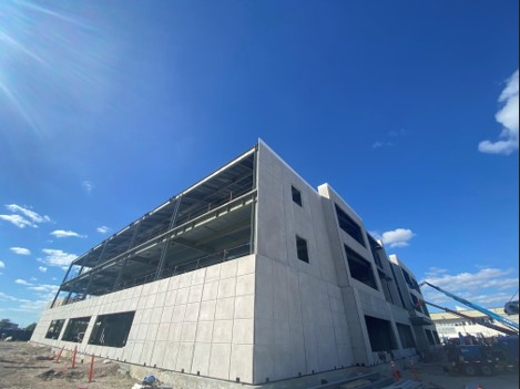 image004 (2) A modern, multi-story building under construction with exposed steel beams and concrete walls, set against a clear blue sky with scattered clouds. Construction equipment and materials are visible on the ground.