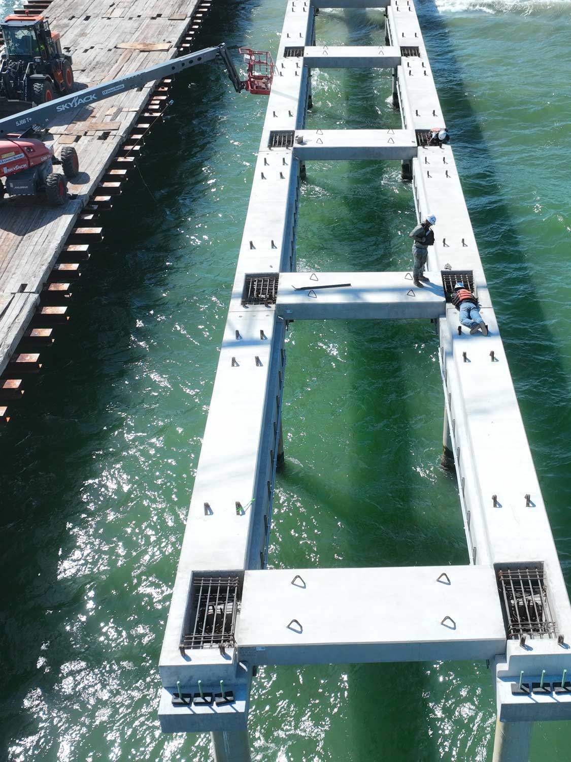 slabbeam-(1) Aerial view of two construction workers standing on a large concrete pier structure over green water, with a nearby crane and equipment on a wooden dock.