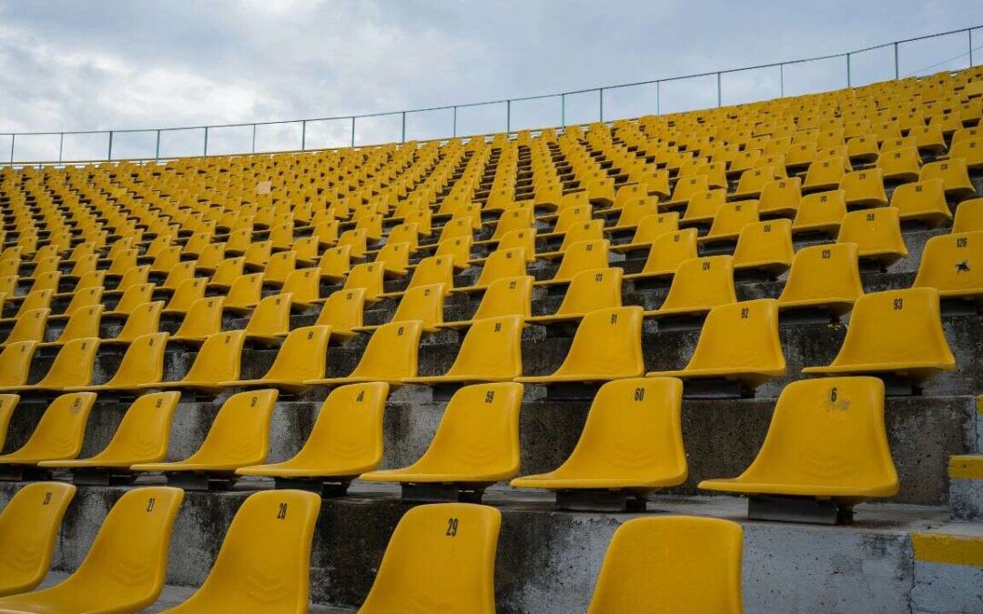 Rows of empty yellow stadium seats are arranged in ascending tiers under a cloudy sky, with some steps visible on the right side.