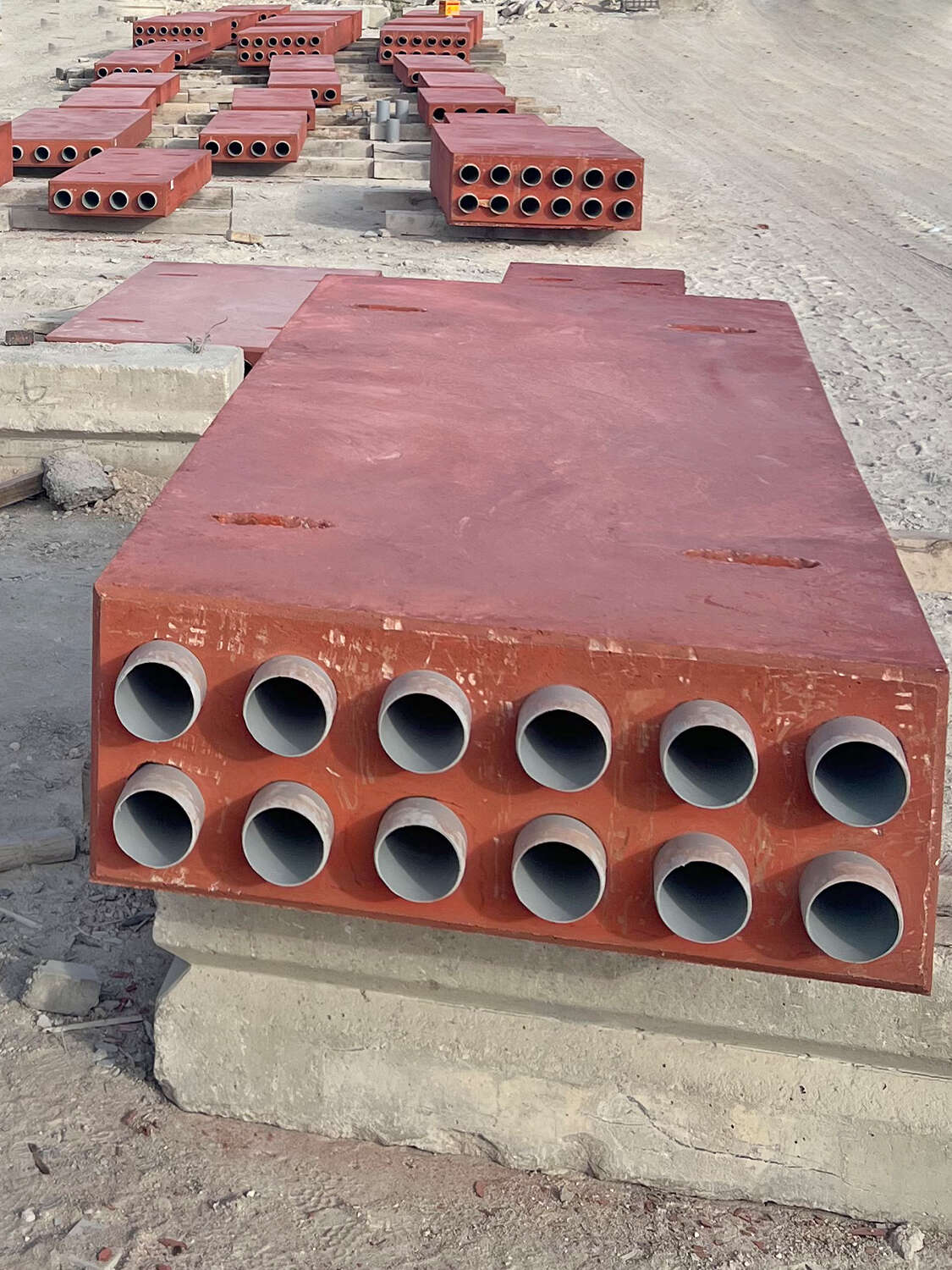 Large rectangular red metal blocks with multiple circular pipes or ducts protruding from one side, arranged on a construction site with concrete and dirt ground. More blocks are visible in the background.
