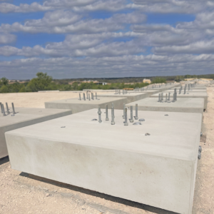 Rows of large, rectangular concrete foundations with metal anchor bolts protruding, set on a construction site under a partly cloudy sky. Trees and landscape are visible in the background.