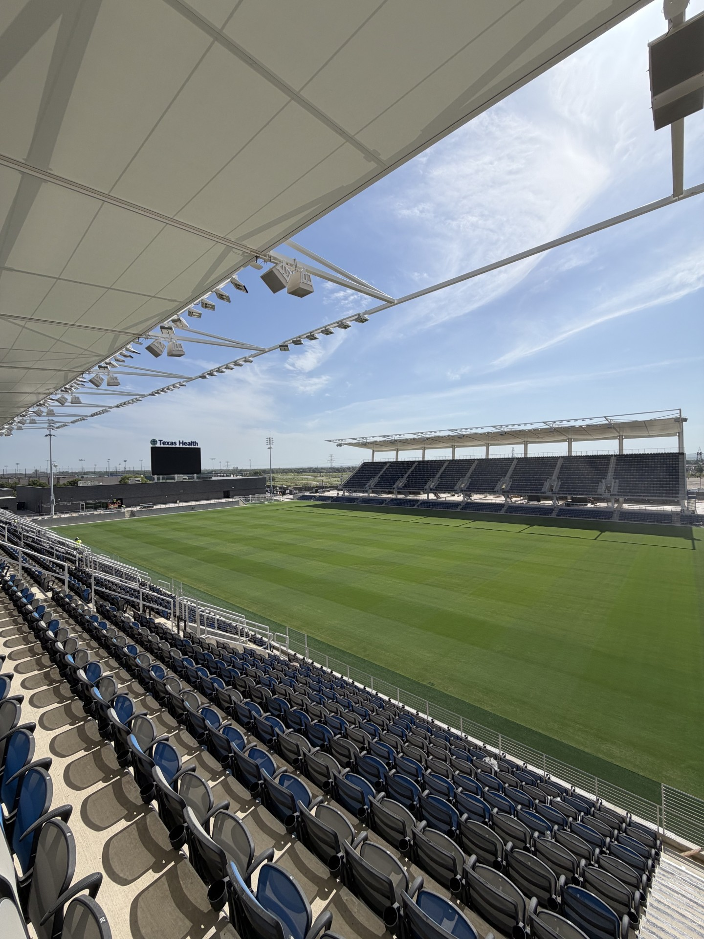 finished1 A wide view of an empty soccer stadium with rows of blue seats, a large green field, and a scoreboard in the distance beneath a clear, sunny sky.