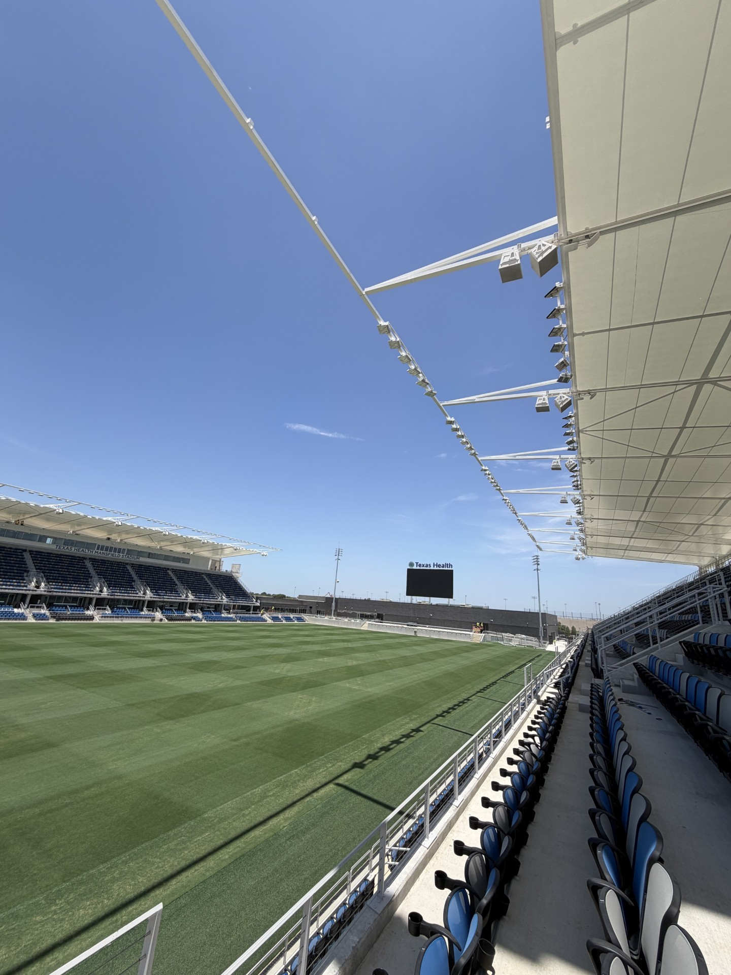 finished2 A wide-angle view of an empty soccer stadium under a clear blue sky, showing green grass, blue and black seating, and a large scoreboard at the far end. The stadium appears modern with a partially covered seating area.